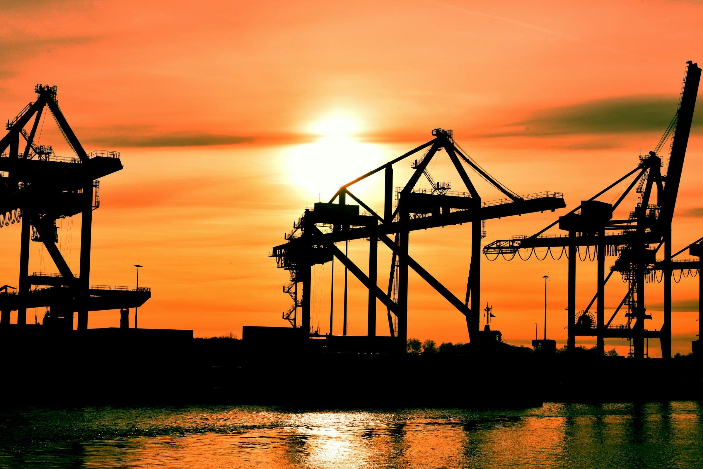 Container cranes silhouetted against an orange sunset over a harbor
